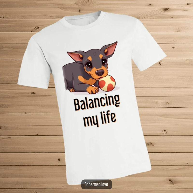 Funny Doberman T-Shirt showing a kawaii doberman puppy focused on balancing a small ball on its nose, a humorous feat.